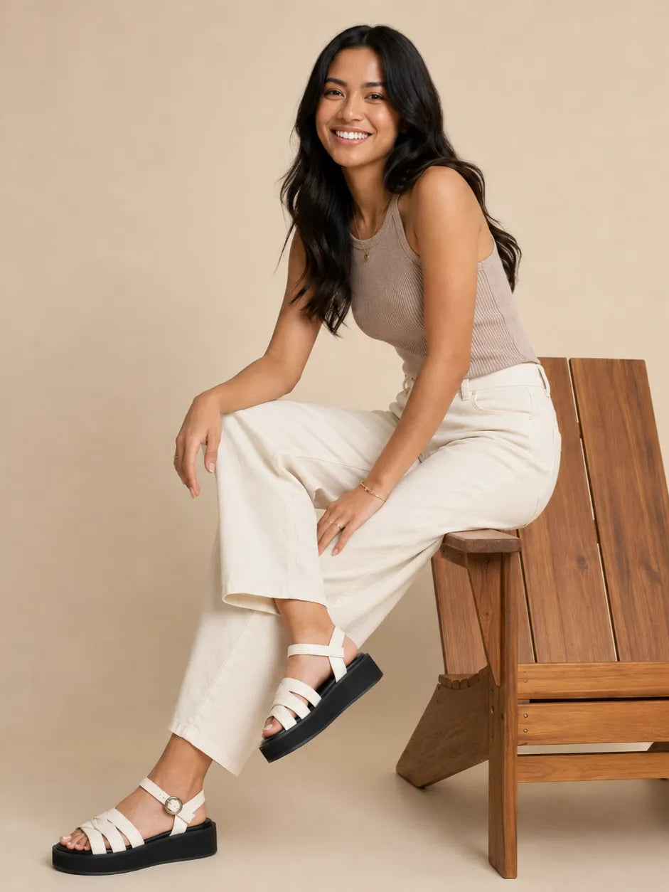 A woman sits on a wooden chair wearing a beige sleeveless top, cream wide-leg cropped pants, and white platform sandals with black soles against a solid beige studio background.