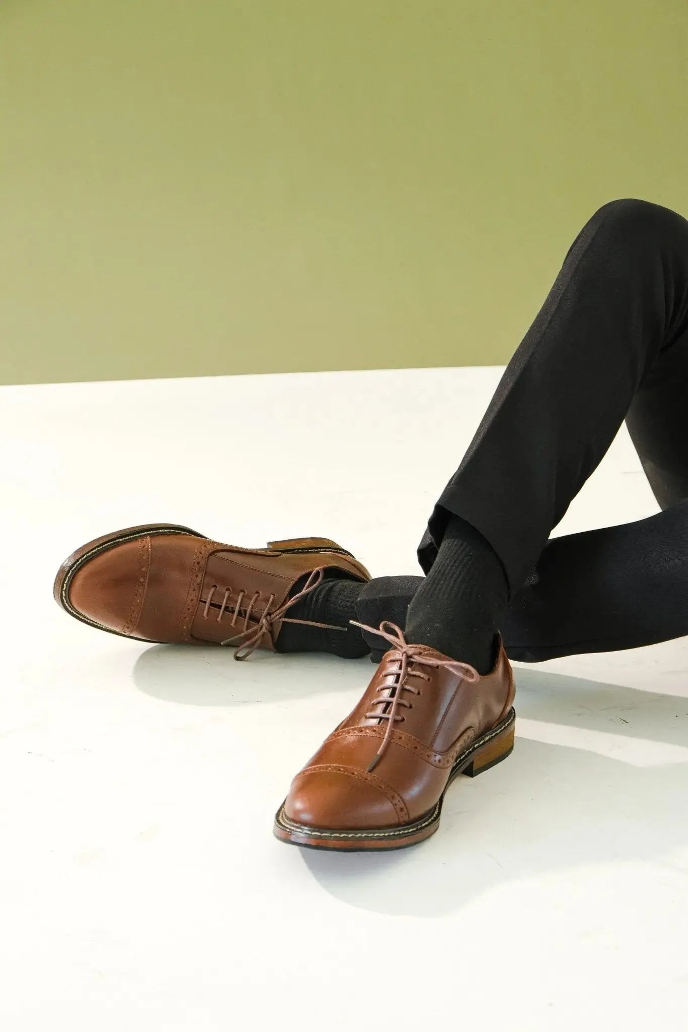 Leather oxford brown leather shoes with holes worn with black pants on a light surface and green wall background
#color_walnut