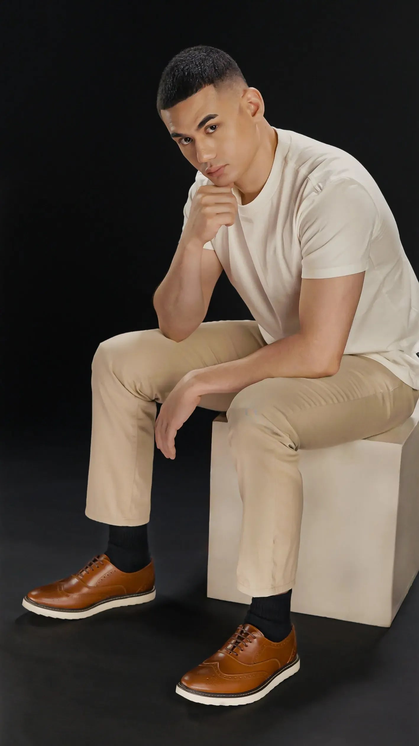 Man sitting on a white cube wearing a beige shirt, beige pants, and tan brown leather shoes with white rubber soles against a black background
#color_tan