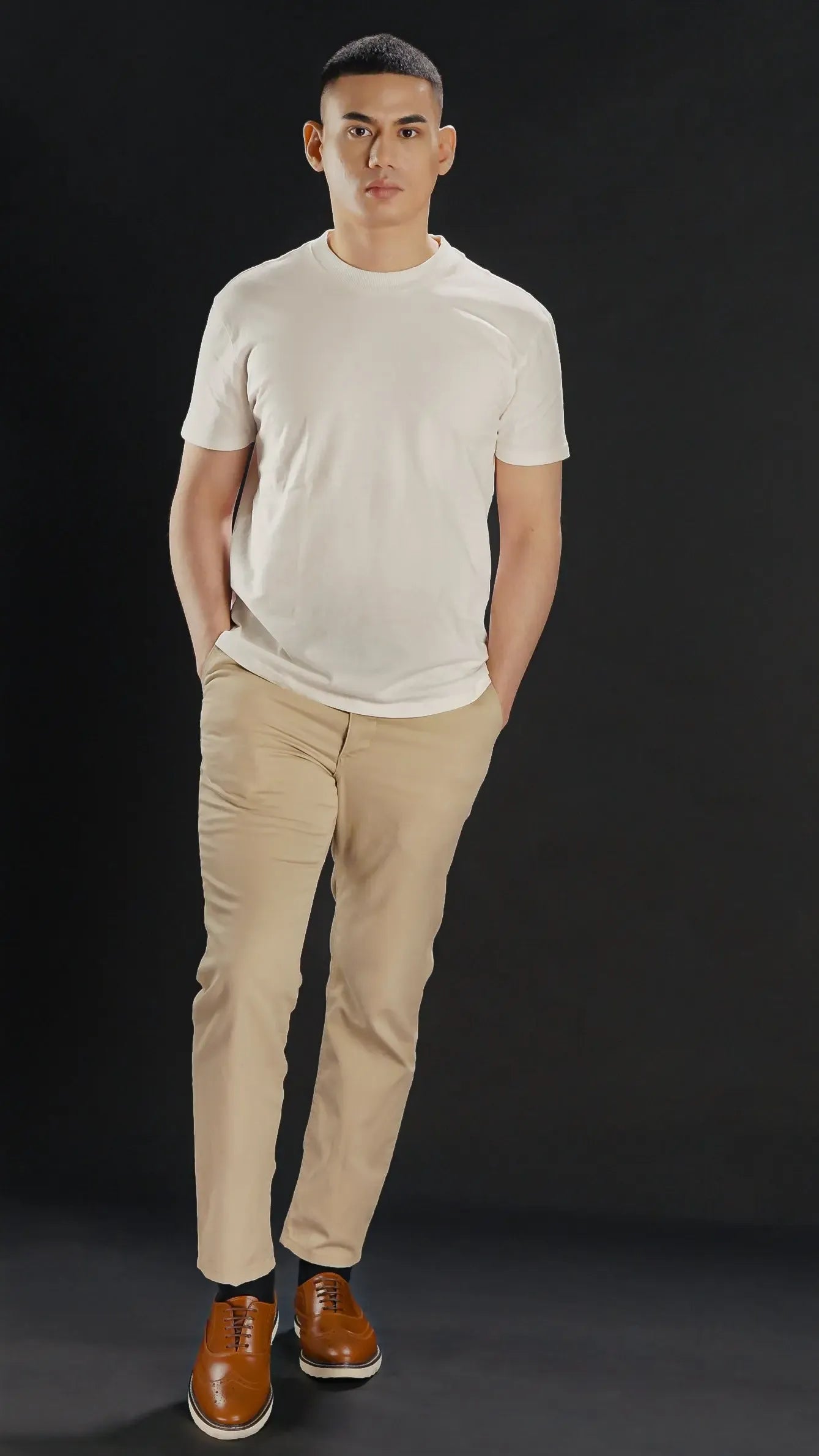 Man wearing a white t-shirt, beige pants and tan brown leather wingtip shoes with white rubber soles against a black background
#color_tan