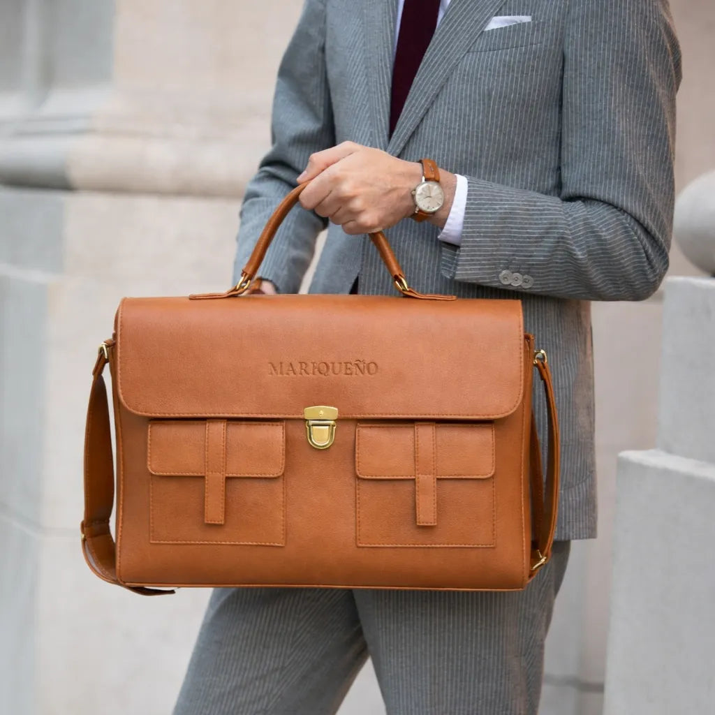 Man holding a brown leather briefcase with 'Marquesao' branding, wearing a gray suit.
#color_tan