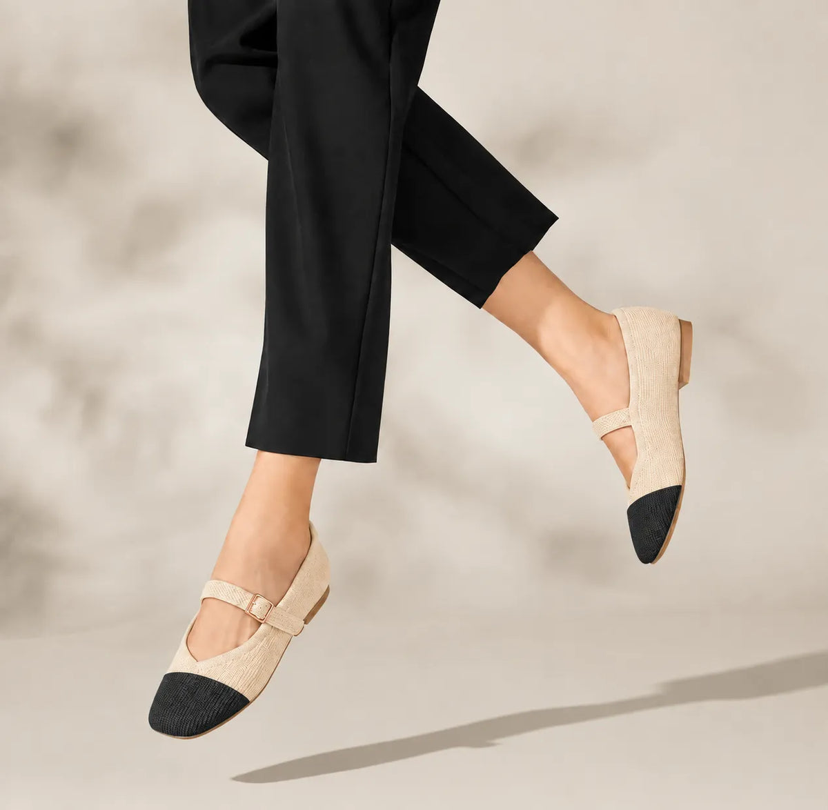 A person wearing black cropped pants and Mary Jane-style flats with beige woven uppers and black toe caps, photographed mid-step against a soft beige studio background.
#color_2 toned