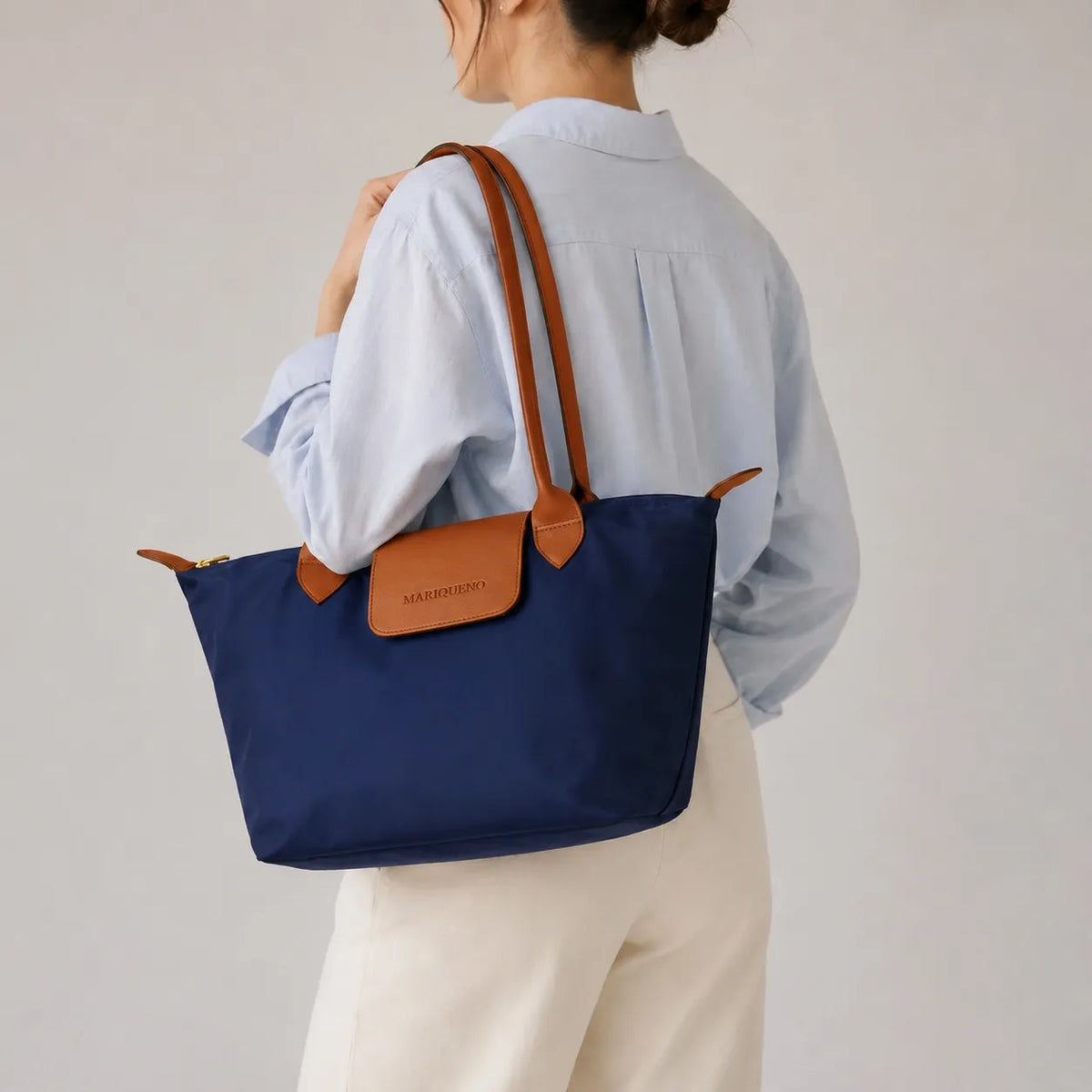 A navy blue tote bag with tan leather handles and a front flap logo patch, carried on the shoulder of a person wearing a light blue shirt and cream pants against a plain light gray background.
#color_navy blue and tan