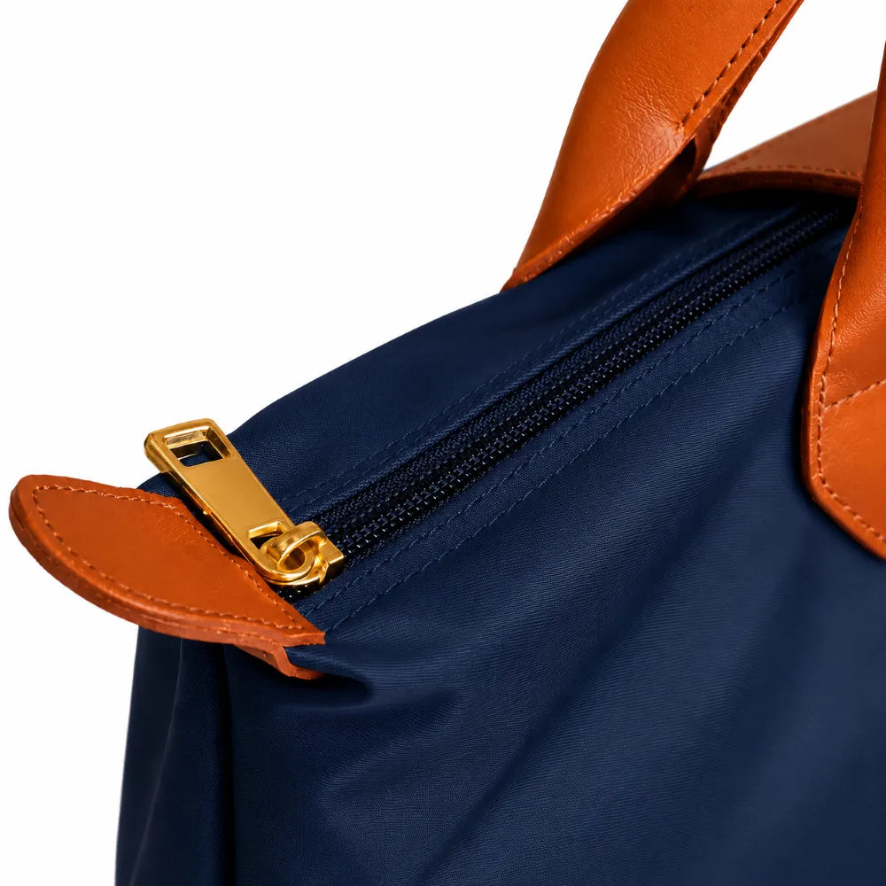 Close-up of a navy blue bag with tan leather trim and a gold-tone zipper pull on a white background.
#color_navy blue and tan