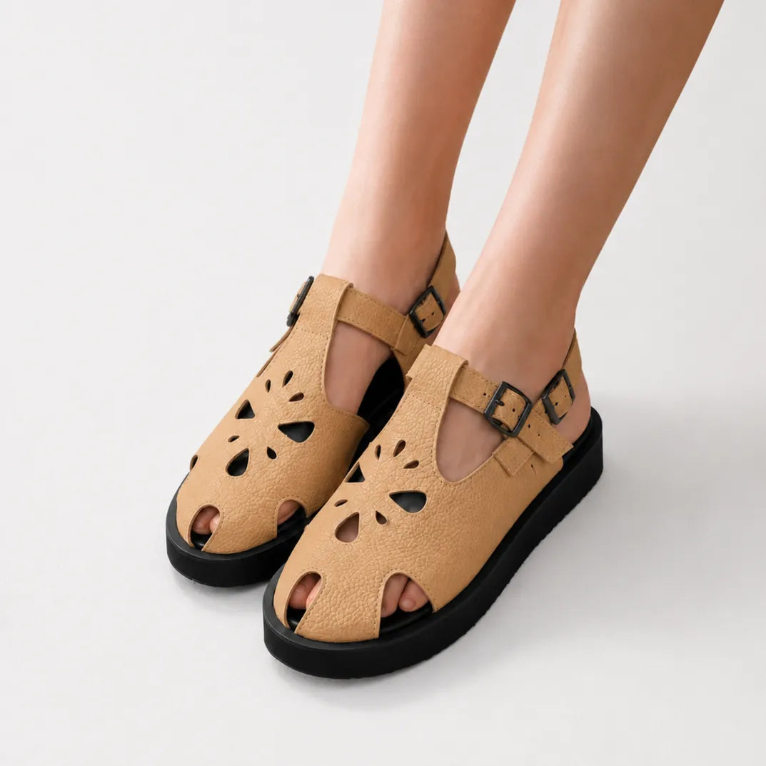 A pair of tan pebbled leather cutout sandals with black soles and adjustable ankle straps, shown on a model's feet against a light gray studio background.
#color_beige
