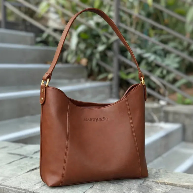 Brown leather handbag with 'MARIQUENO' branding on a stone surface with blurred greenery in the background.
#color_choco brown
