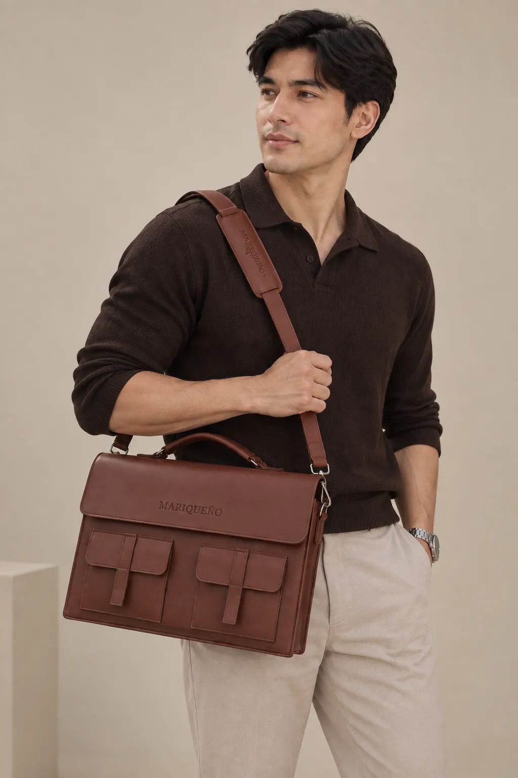  man models a brown leather briefcase with a matching shoulder strap. He wears a dark brown long-sleeve knit polo shirt and beige trousers. The briefcase features two front utility pockets and the brand name "MARIQUEÑO" debossed on the flap.