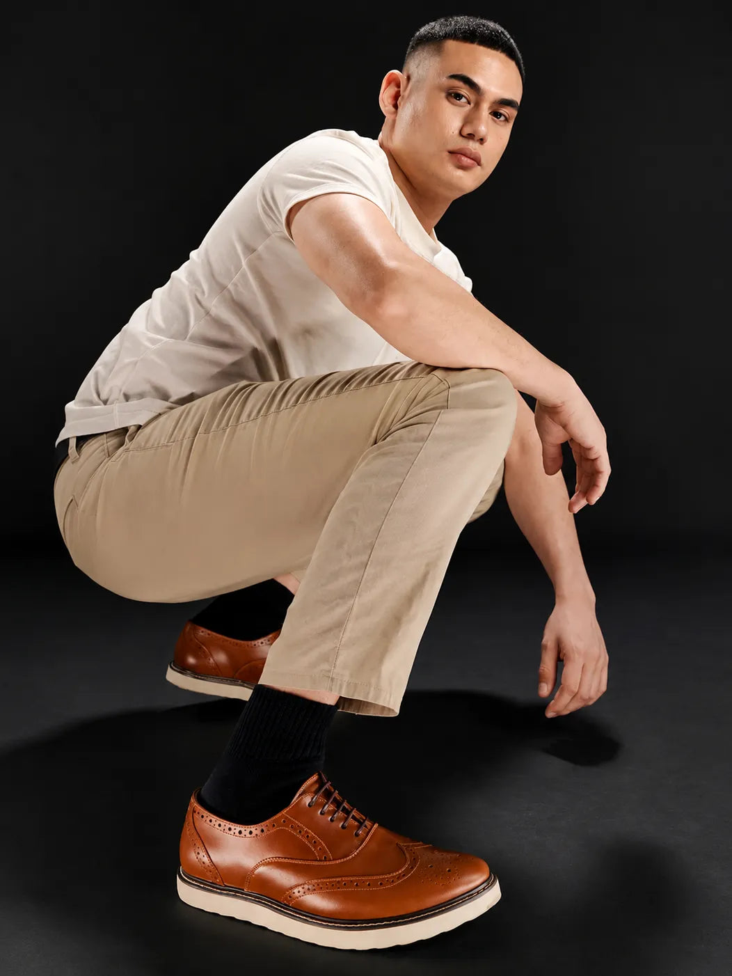 A man crouching in a studio setting, wearing a white t-shirt, beige trousers, black socks, and brown brogue oxford shoes against a dark background.