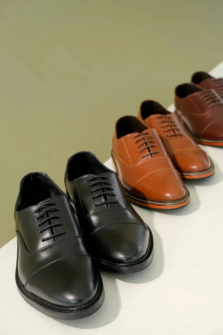 Four men's leather oxford shoes arranged in a row on a white surface against a muted green background. The front pair is black, and the pairs behind are tan and brown.