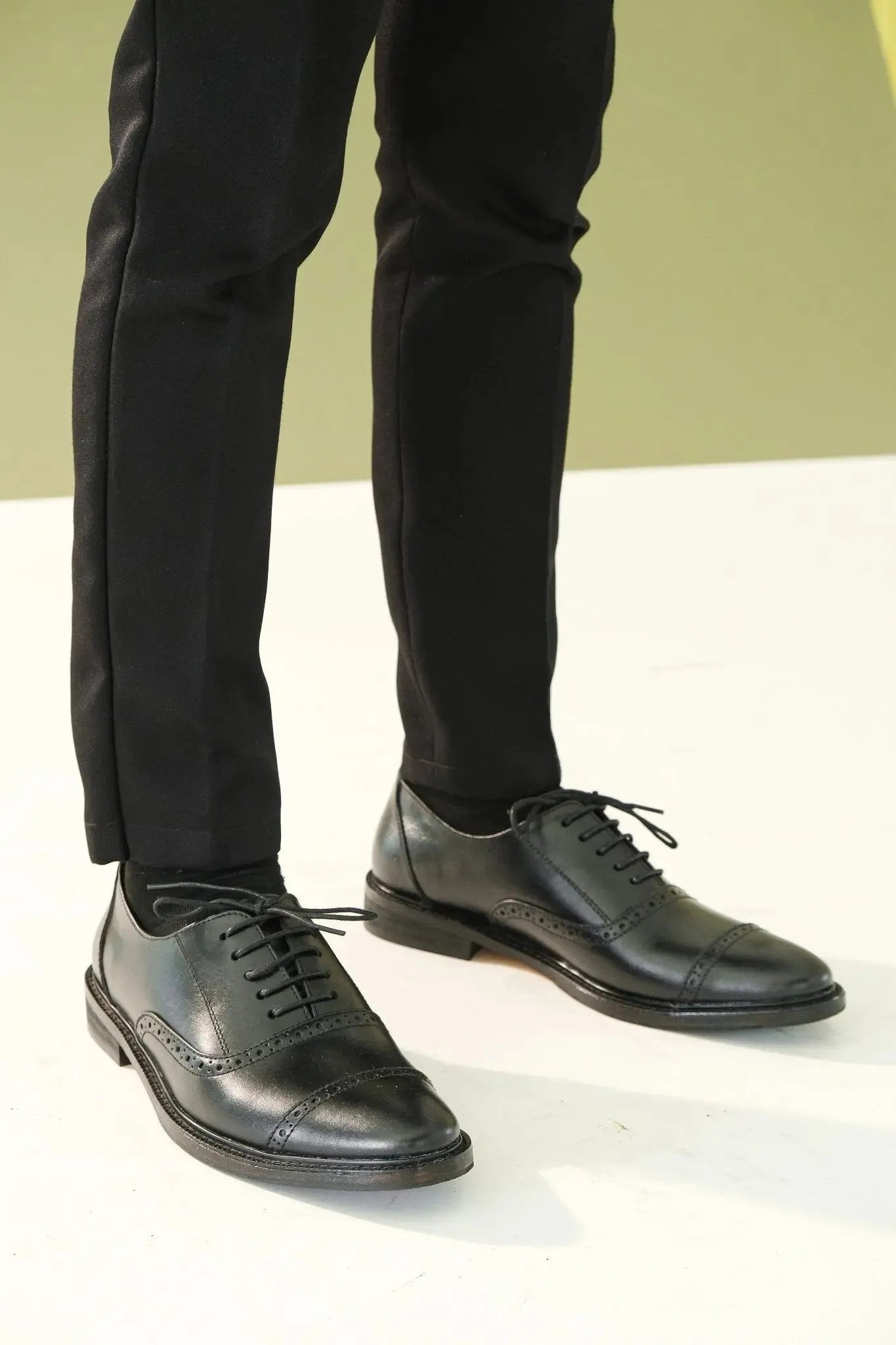 Black oxford shoes with holes and pants on a white surface with a light green background
#color_black