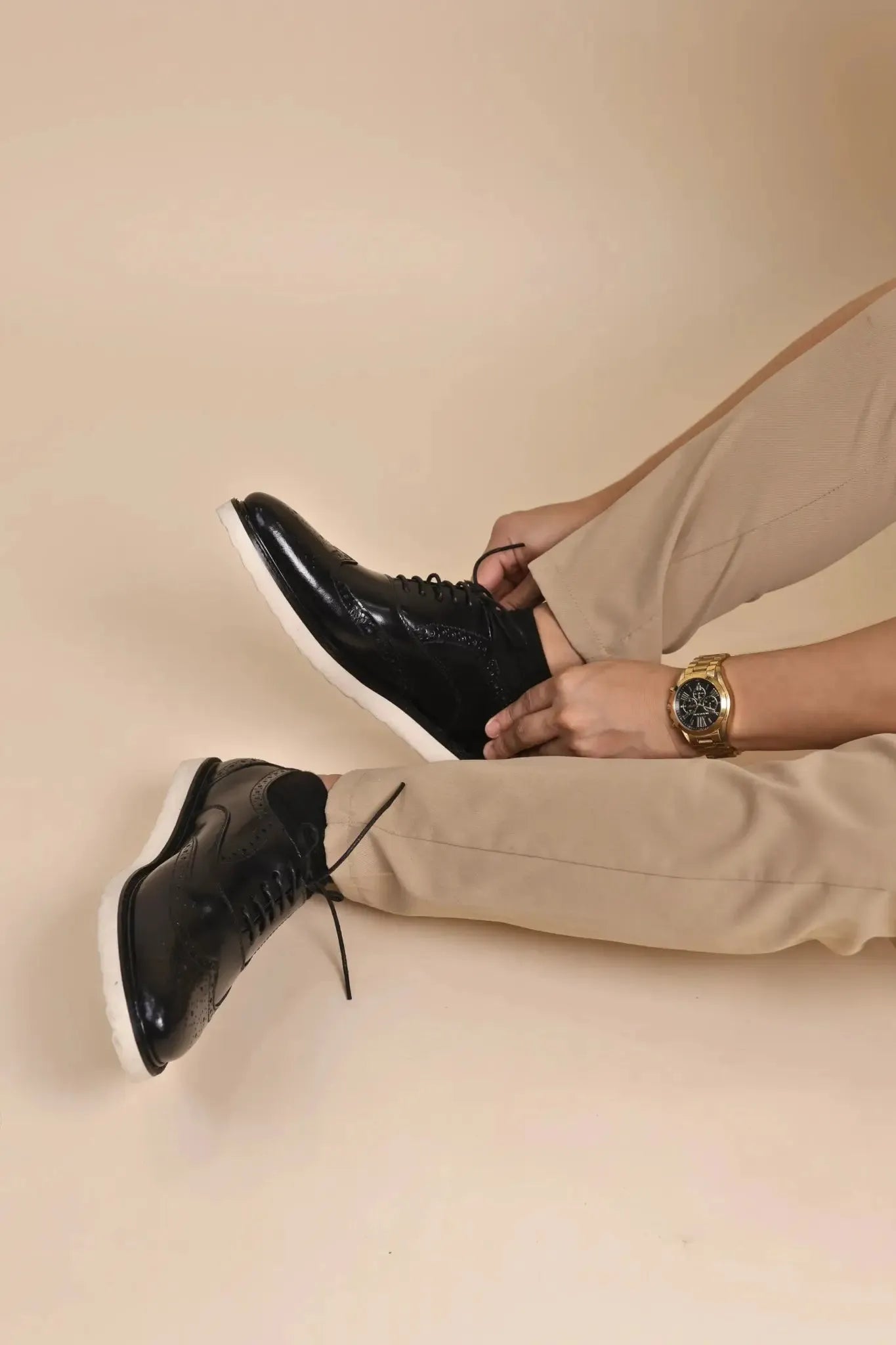 Person wearing black leather wingtip shoes with white rubber soles and beige pants with a watch on a beige background
#color_black