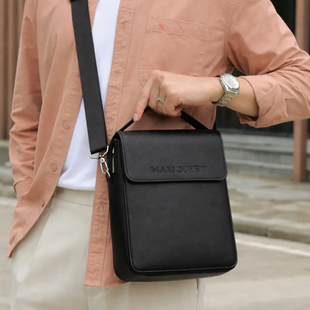 Person holding a black crossbody bag with 'Marqueno' branding, wearing a light brown jacket.
#color_black