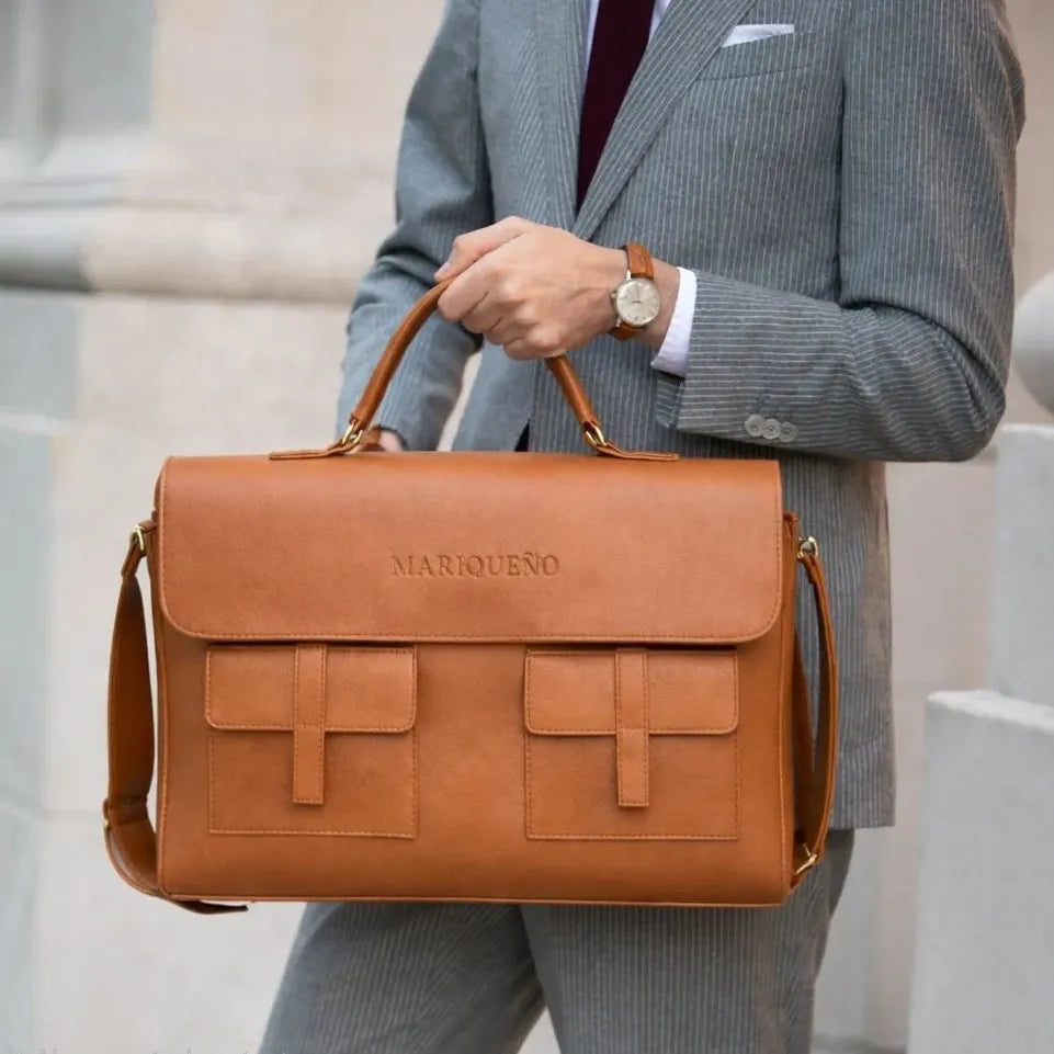 Man holding a brown leather briefcase with 'Marquesao' branding, wearing a gray suit.
#color_tan