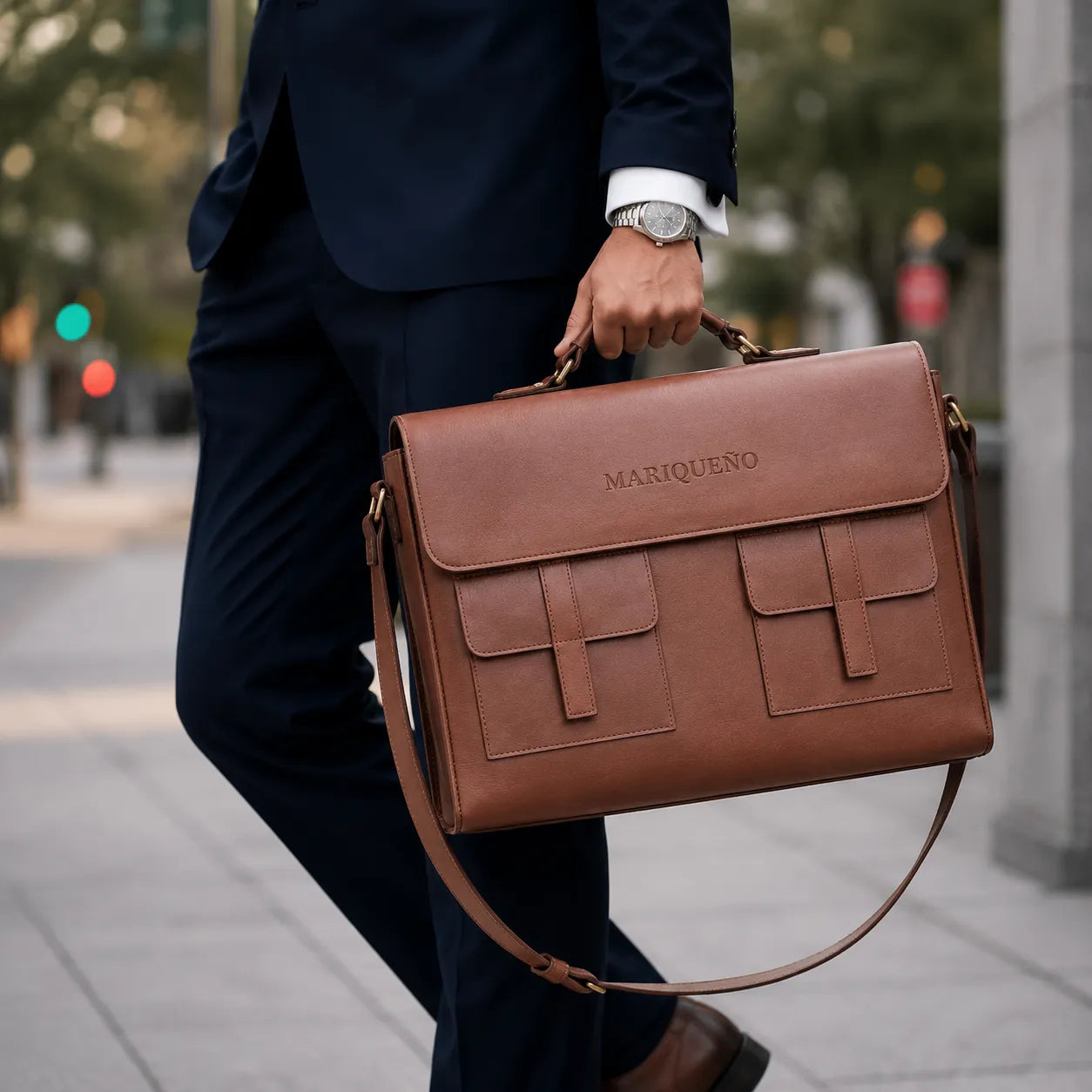 Person holding a brown leather briefcase with 'MARIQUENO' branding on a city street.
#color_walnut