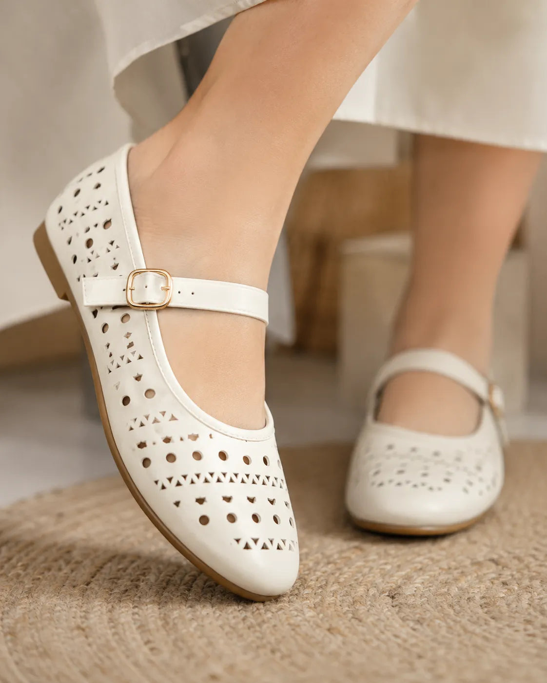 A pair of off-white Mary Jane shoes with laser-cut circular and geometric patterns, featuring a single strap with a small gold buckle. One shoe is shown being worn on a foot, while the other is placed on a woven jute rug.
#color_white