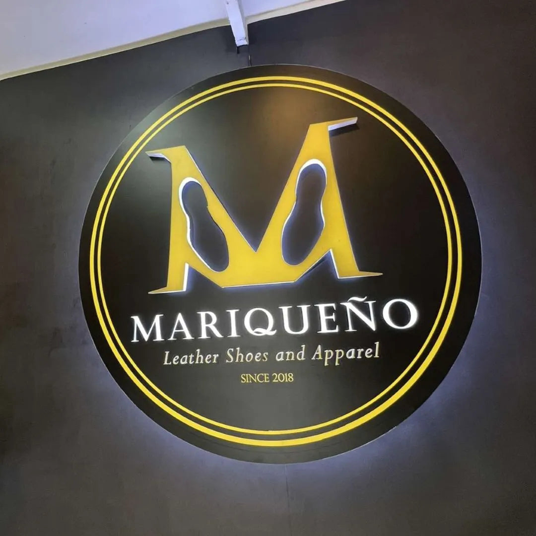 A circular black and yellow illuminated sign on a dark wall featuring a large stylized 'M' logo and the text 'MARIQUEÑO Leather Shoes and Apparel SINCE 2018'.
