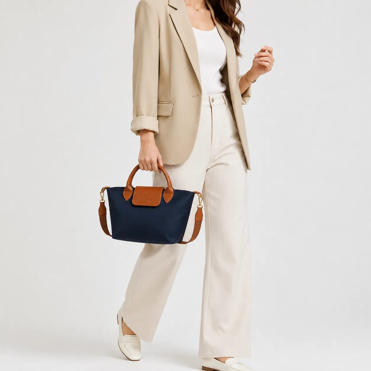 A woman models a navy blue canvas handbag with brown leather handles and trim, styled with a beige blazer, white top, and cream wide-leg trousers against a light gray studio background.
#color_navy blue