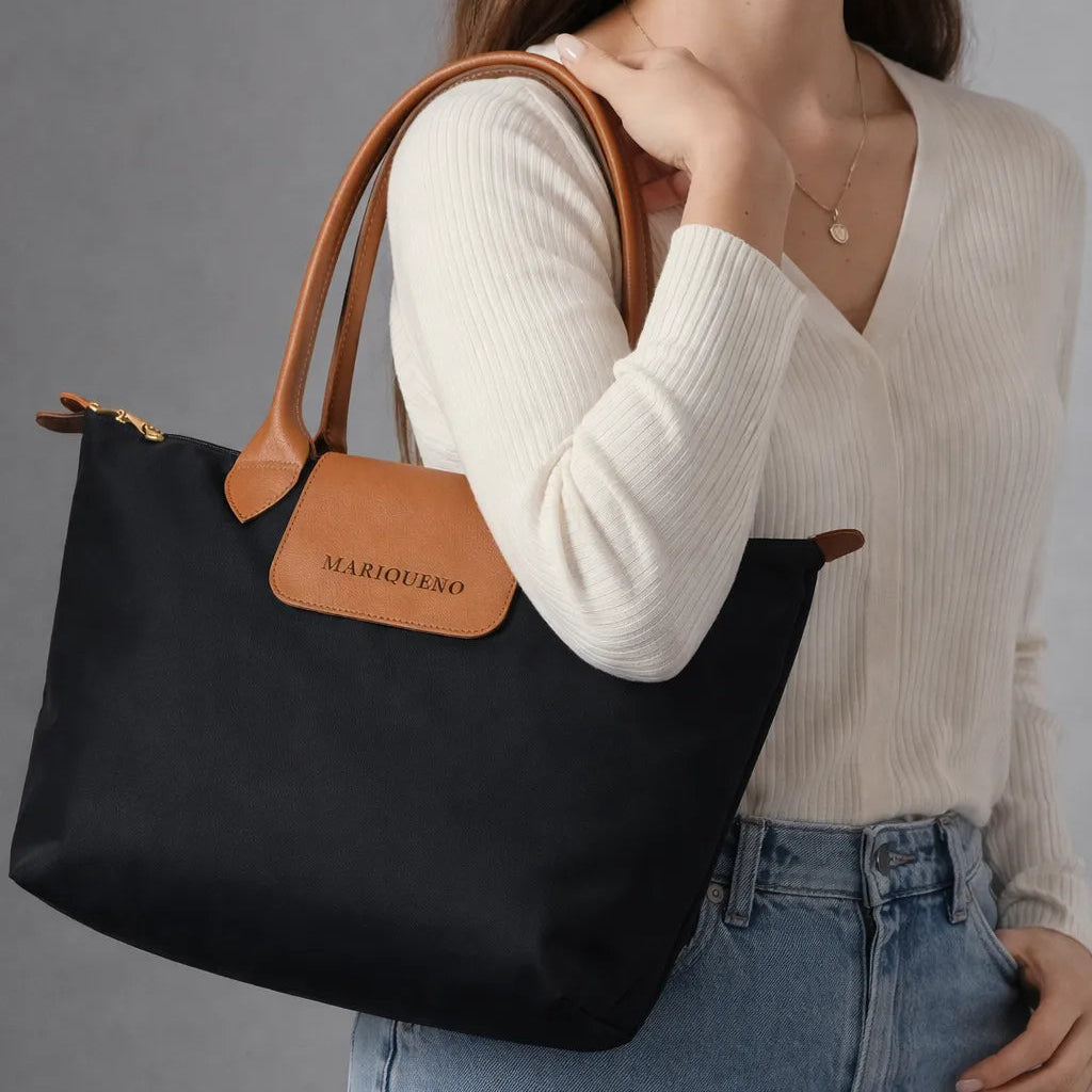 Person holding a black tote bag with brown handles and a visible brand label against a gray background
#color_black