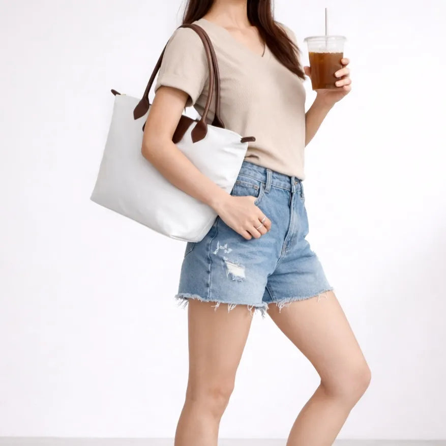 Person wearing a beige top and denim shorts, holding a white tote bag and a drink.
#color_white
