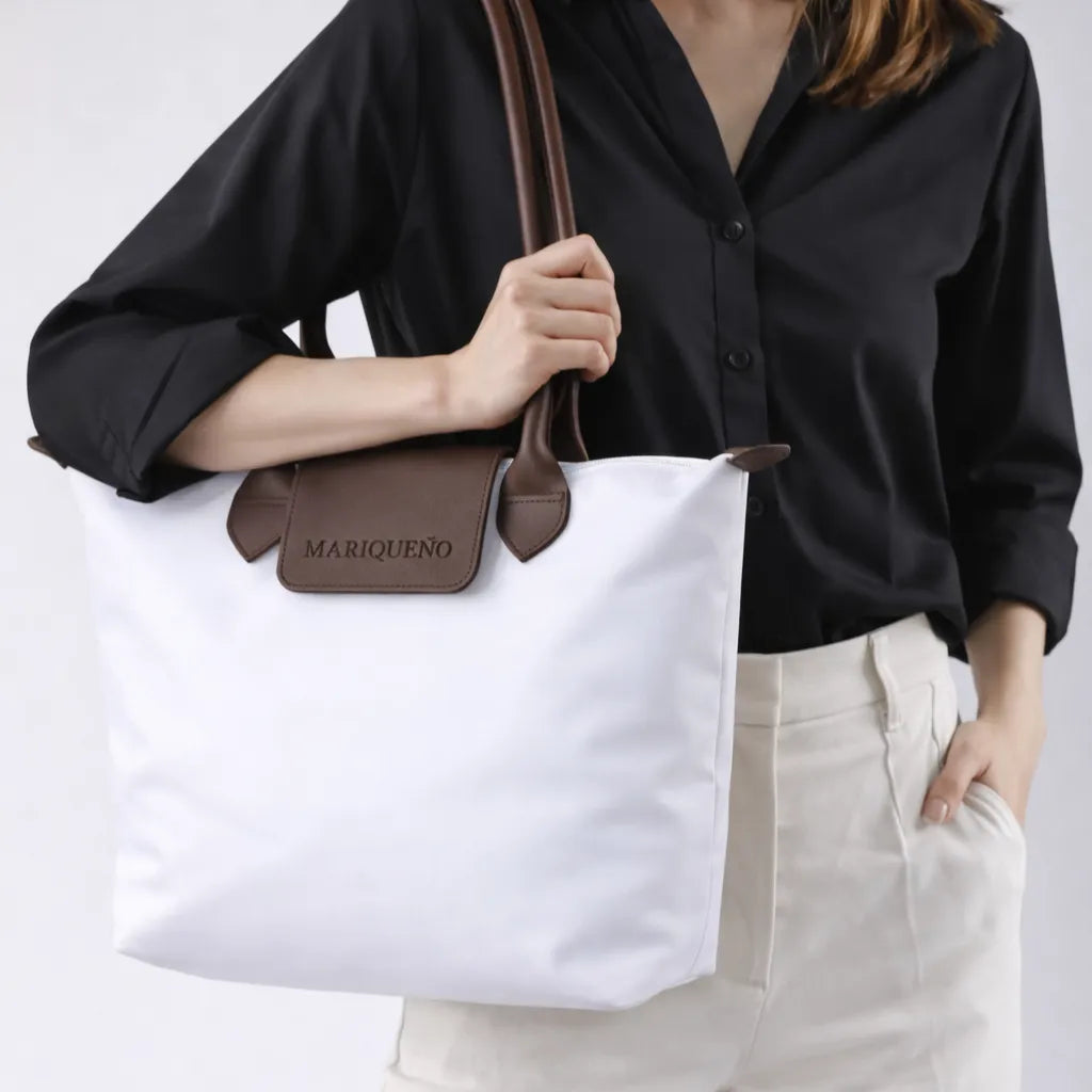 Person holding a white tote bag with brown handle and 'MARIQUENO' branding.
#color_white