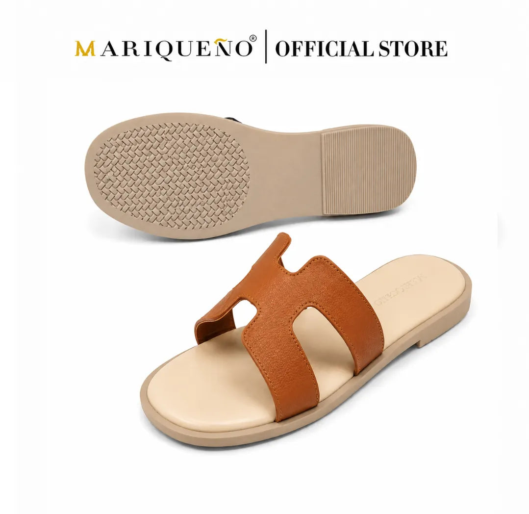 A pair of MARIQUEÑO slide sandals shown on a white background, with one sandal displaying the tan textured outsole and the other showing the side profile. The sandals feature a brown leather H-strap and a beige footbed.
#color_tan