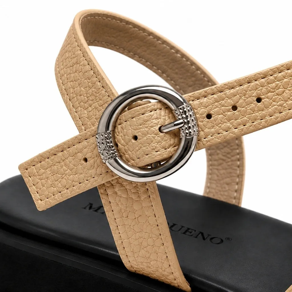 A close-up of a beige textured leather sandal strap with a silver-tone circular buckle and rhinestone accents, resting on a black shoebox. The brand name 'MARIQUENO.
#color_beige
