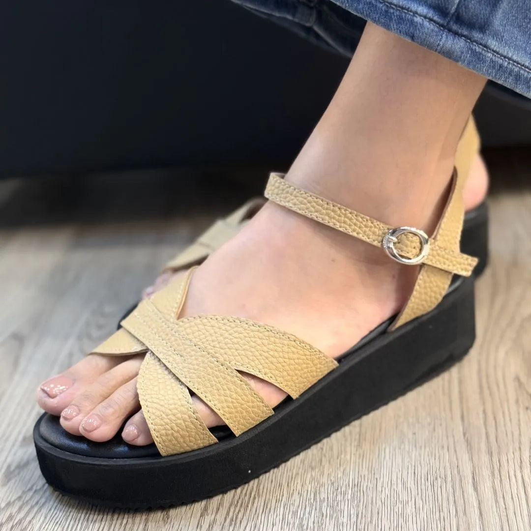 A close-up of a person's foot wearing a tan textured wedge sandal with crisscross straps and a black platform sole, standing on a light wood floor.
#color_beige